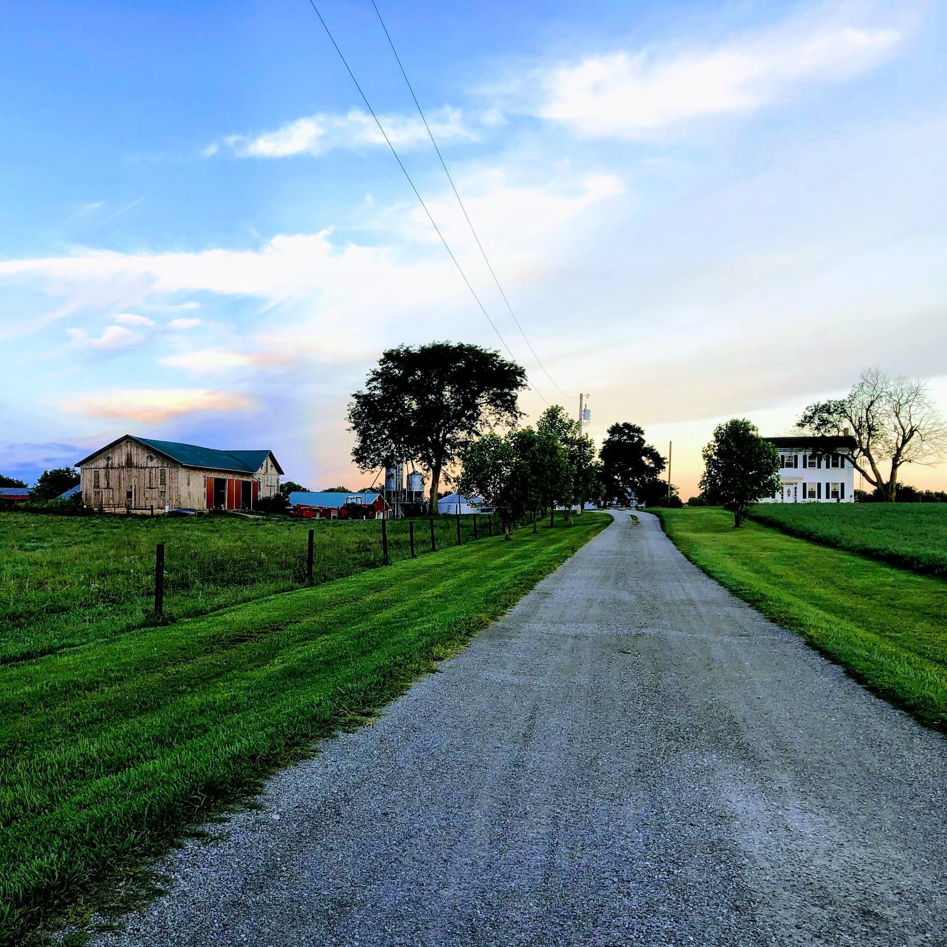 Gravel road leading to a rural farmhouse and barn, surrounded by lush green grass and trees under a colorful sunset sky. Ideal for showcasing serene countryside landscapes and agricultural settings.