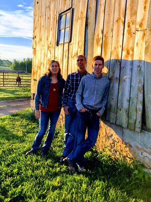 Three individuals pose casually against a rustic wooden barn, surrounded by lush green grass and a clear blue sky. The scene captures a warm, sunny day on a farm, highlighting a sense of camaraderie and outdoor lifestyle.
