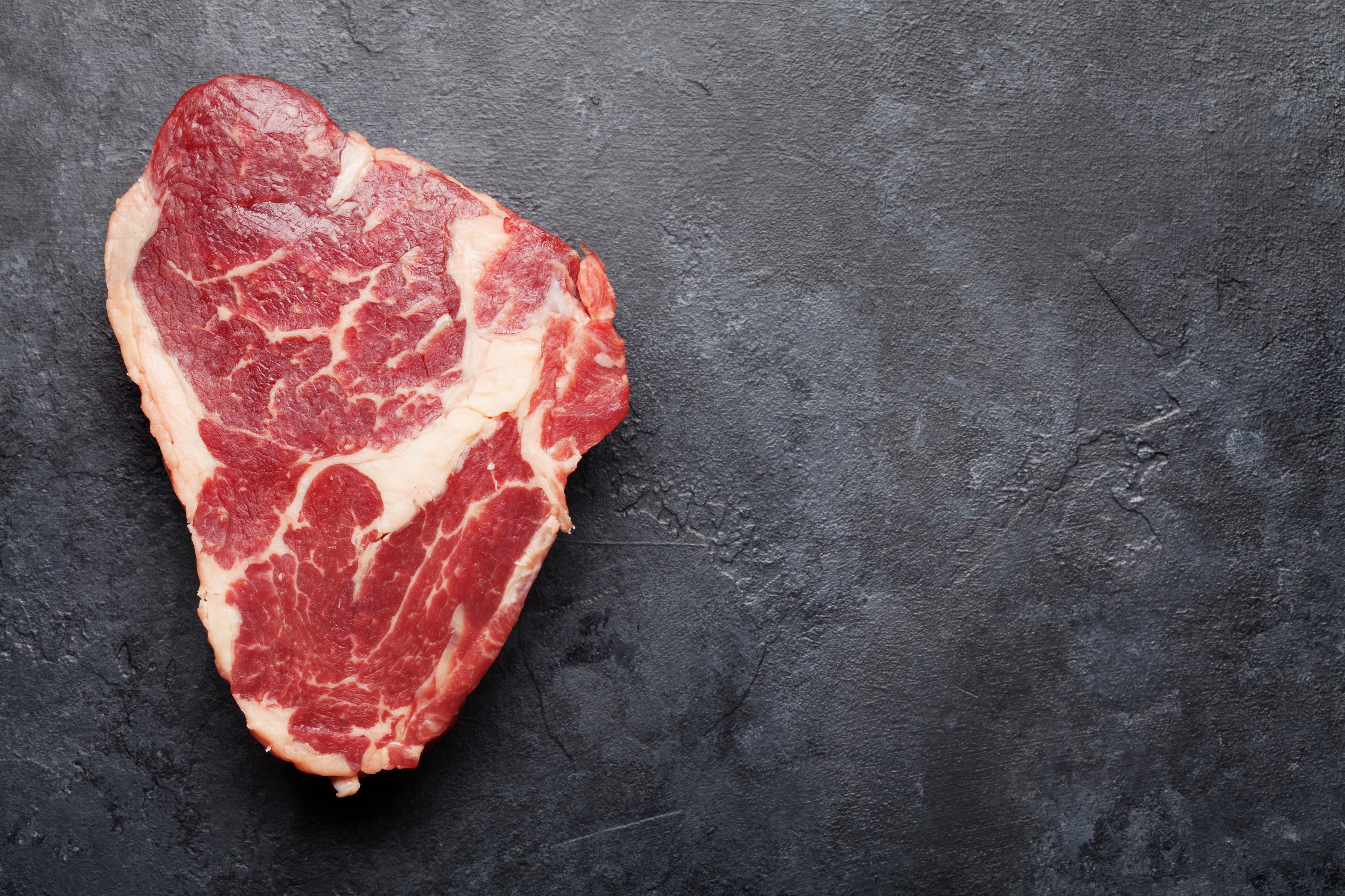 Ribeye raw beef steak Fresh, raw ribeye steak on a dark textured surface, showcasing marbling and rich red color, ideal for grilling or slow cooking.