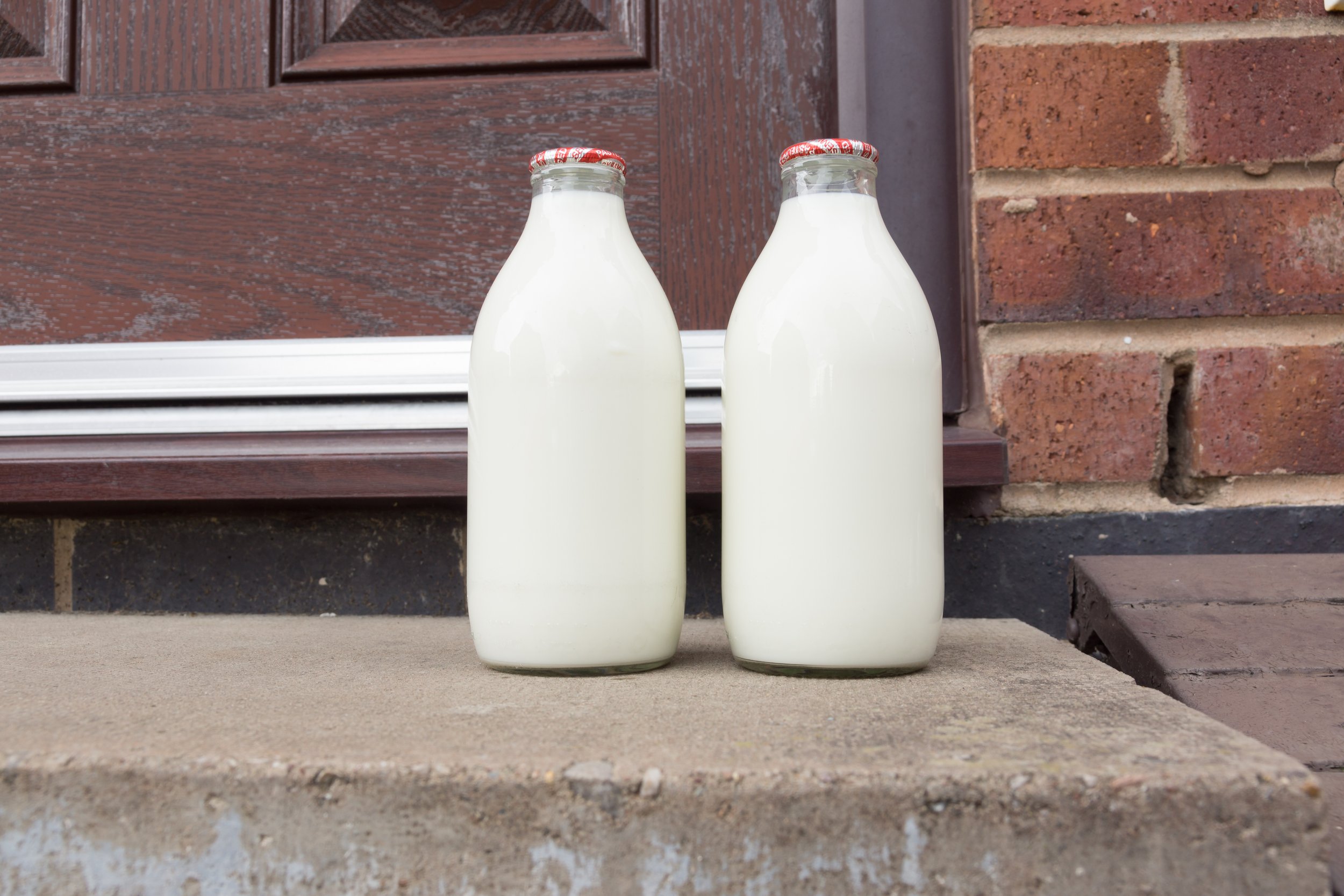 Fresh milk in recyclable glass bottles delivered to the door by a traditional milk man Two glass bottles of fresh milk placed on a concrete step in front of a wooden door, showcasing a rustic and home-delivery feel.