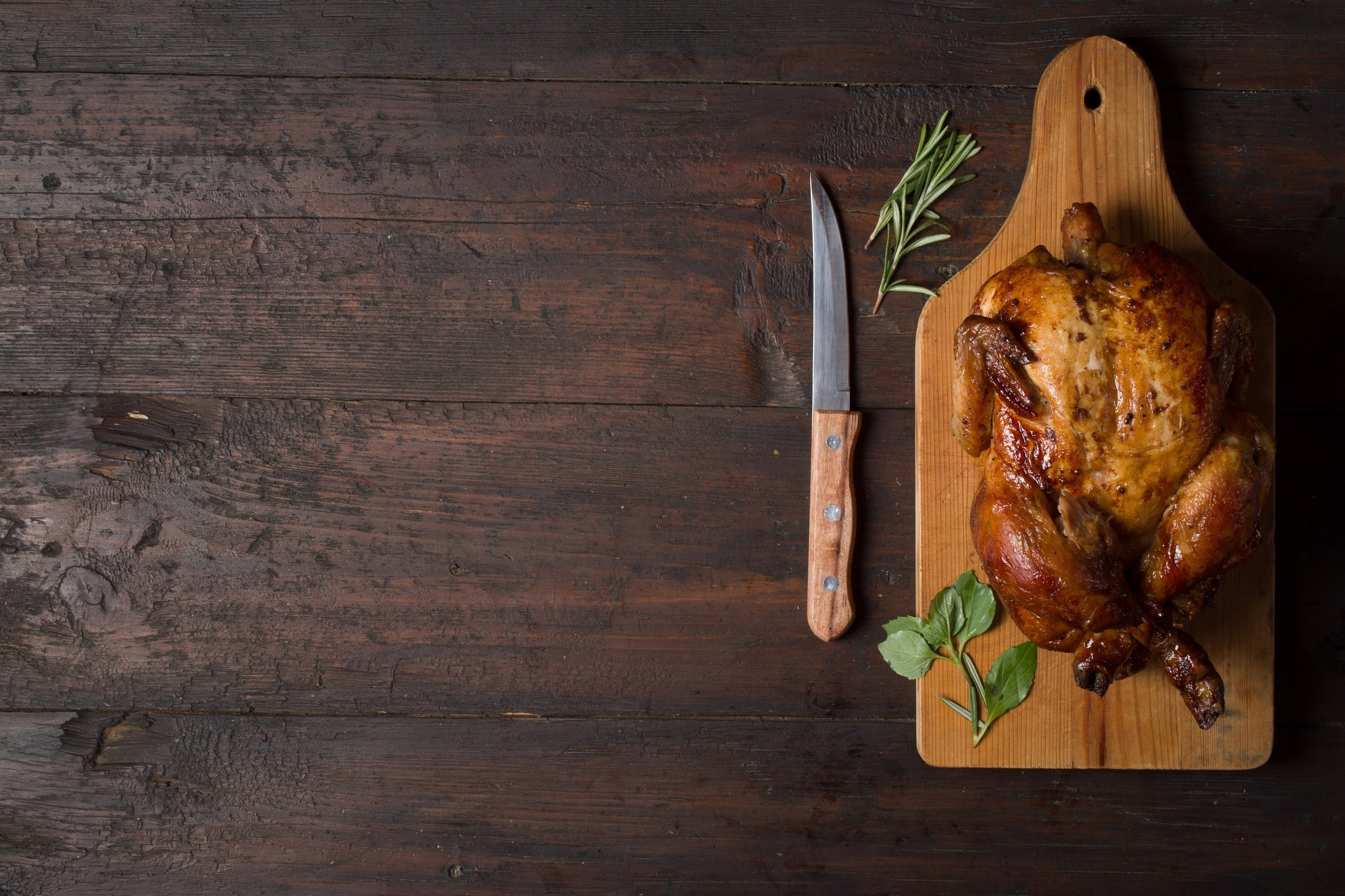 chickenorturkey_roast Roasted chicken on a wooden cutting board, accompanied by a knife and fresh herbs, set against a rustic wooden background.