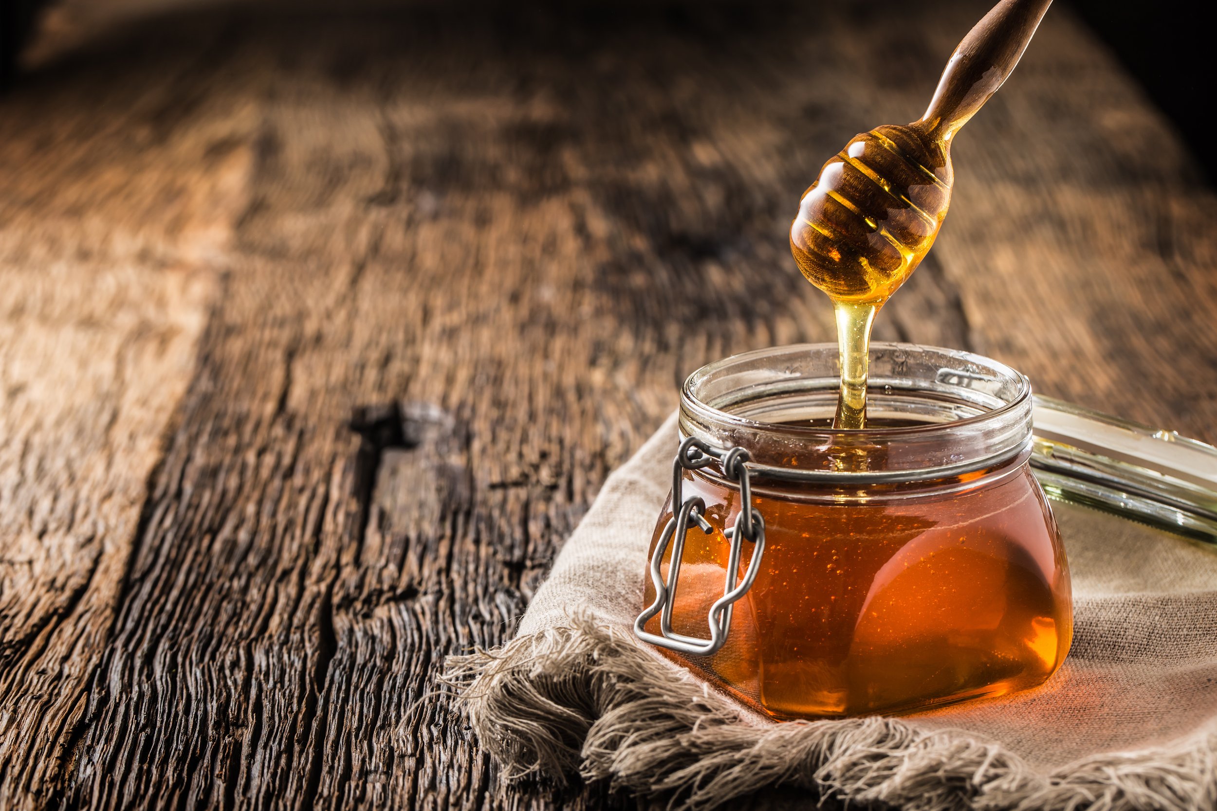 Honey jar with dipper on old woodn table. Glass jar of golden honey with a wooden dipper, placed on a rustic wooden surface, surrounded by a linen cloth. The honey is dripping from the dipper, showcasing its thick, viscous texture.